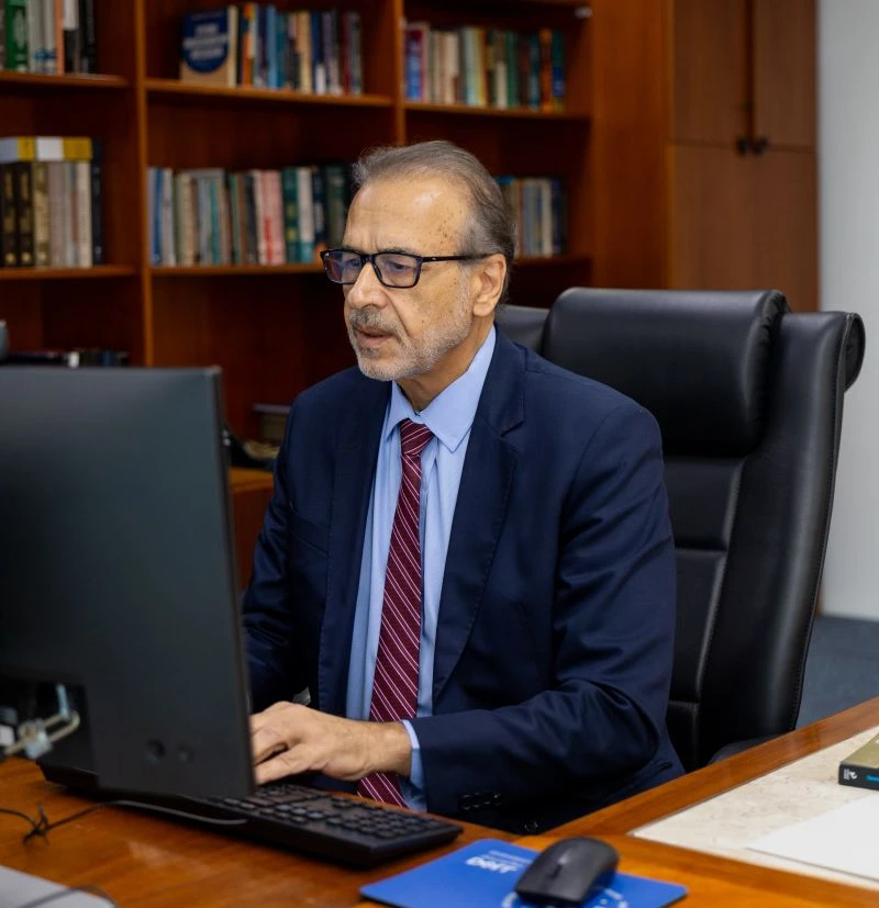 Dr. Ali Mazloum em seu gabinete, ao computador, diante da estante de obras jurídicas